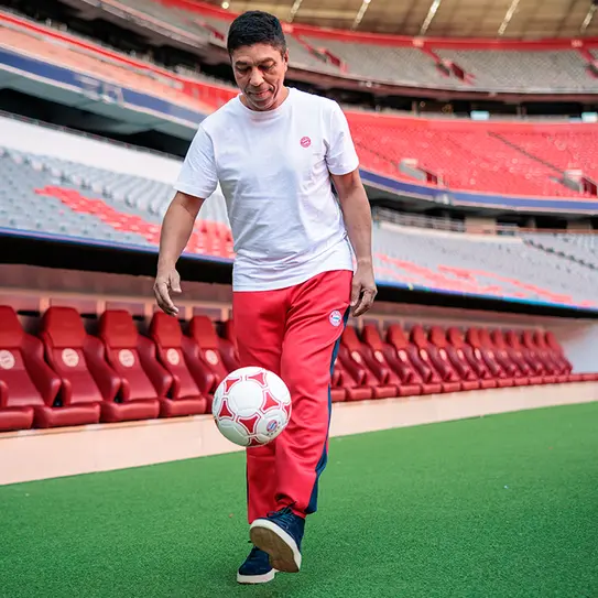Un bărbat poartă un tricou alb și pantalonii roșii în timp ce driblează o minge de fotbal pe terenul unui stadion. În fundal se poate observa un rând de scaune roșii. Stadionul este gol.