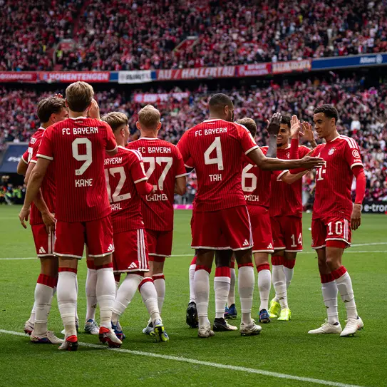 Jucătorii echipei FC Bayern München, îmbrăcați în tricouri roșii, se strâng în jurul unui coleg pe terenul de fotbal, celebrând un moment împreună în fața unui public numeros. Atmosfera este plină de energie și entuziasm.