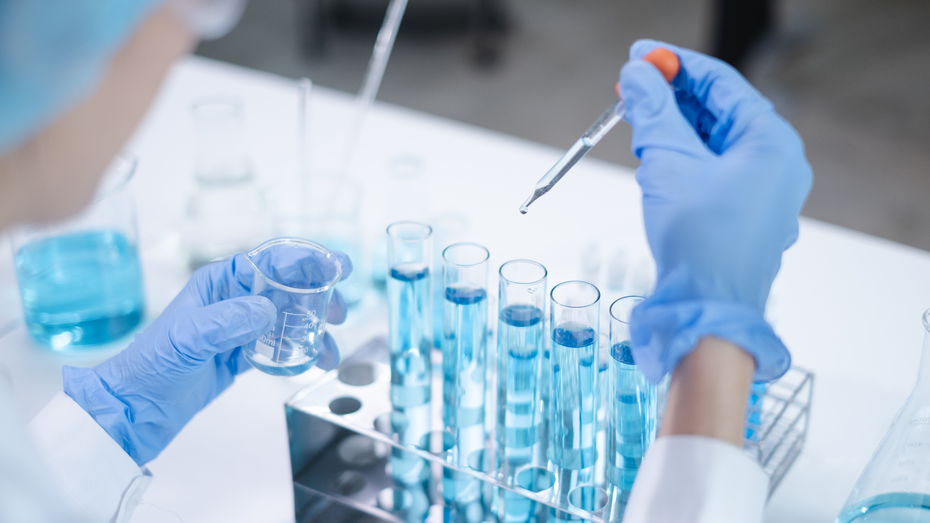 O persoană îmbrăcată în halat de laborator și mănuși, care folosește o pipetă pentru a adăuga un lichid albastru într-un tub de test. În fața sa, se află mai multe tuburi de test și un pahar cu un lichid similar.