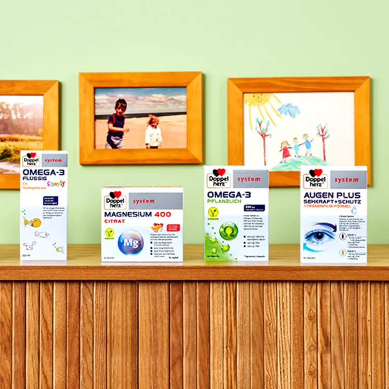 Produse de sănătate expuse pe un raft de lemn, inclusiv cutii pentru suplimentele Omega-3, Magneziu 400 Citrati și Augen Plus. Fundalul include poze de familie și desene realizate de copii, într-un decor de cameră prietenoasă.