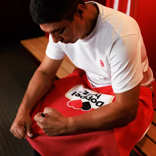 Un bărbat îmbrăcat în tricou alb, stând într-o cabină de vestiar, se uită cu atenție la un prosop roșu cu un design pe el. El își folosește o mână pentru a-l fixa, concentrându-se pe detalii.
