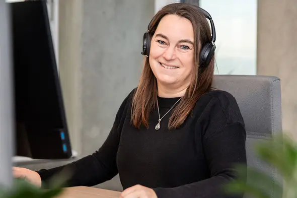 O femeie zâmbitoare, cu căști audio, stă la birou în fața unui monitor. Poartă un pulover negru și are părul lung, desfăcut. Fundalul este simplu, cu elemente de birou.