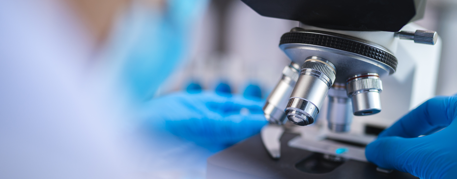 Imaginea prezintă un microscop de laborator, cu detalii asupra obiectivelor și unei mâini îmbrăcate în mănuși albastre care ajustează un specimen pe platou. Fundalul este neclar, sugerând un mediu de cercetare.