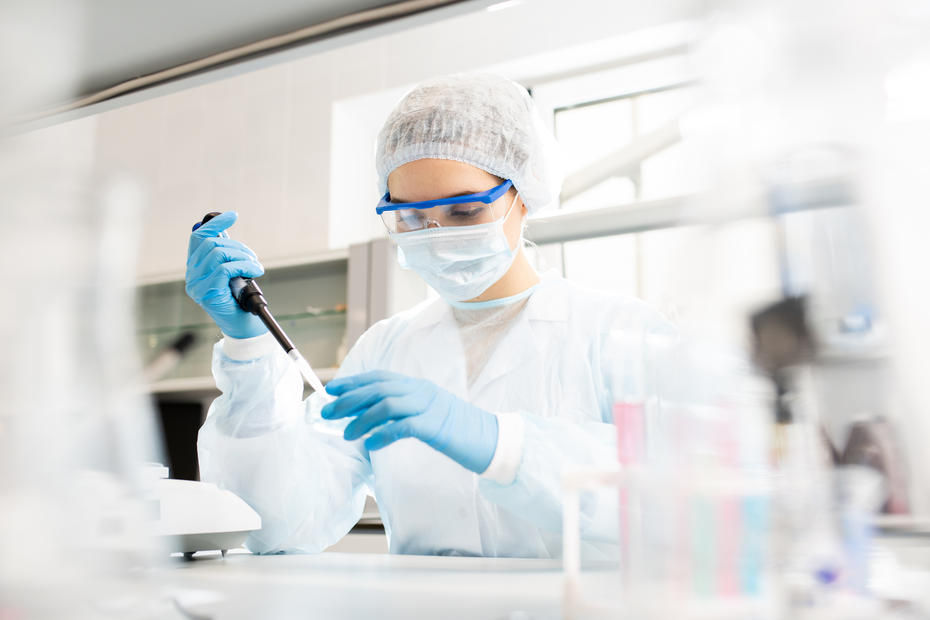 O persoană îmbrăcată în echipament de protecție, inclusiv halat, mănuși și ochelari de protecție, folosește o micropipetă într-un laborator. Se concentrează asupra unui eșantion, într-o atmosferă de cercetare științifică.